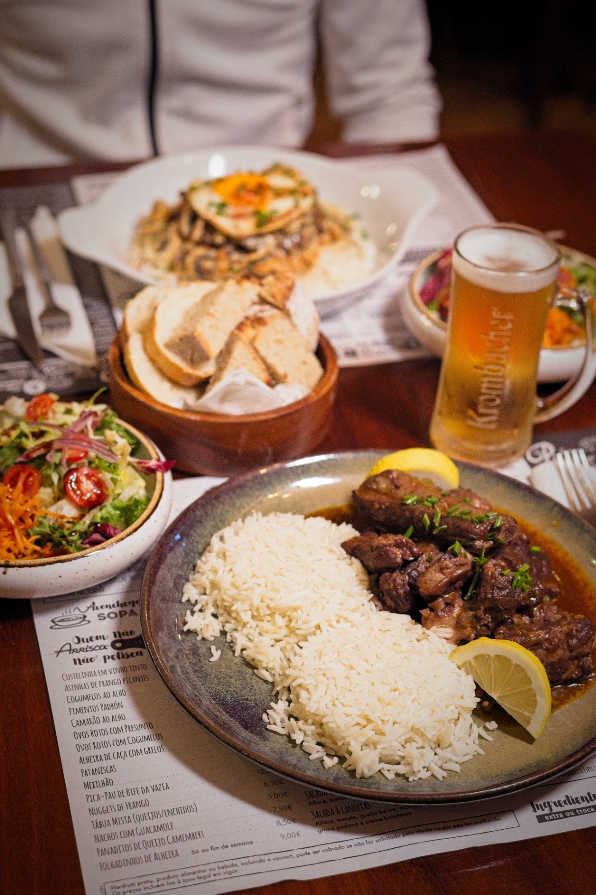 Mesa com carne estufada, arroz, pão, salada e cerveja Krombacher.
